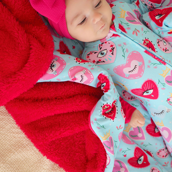 Baby in a colorful outfit with heart patterns lying on a red blanket.