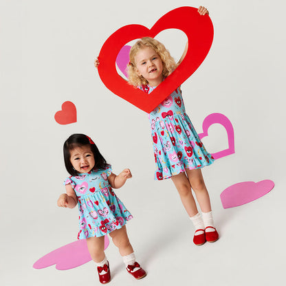 Two children in matching heart-themed dresses standing behind a large red heart cutout on a light gray background.