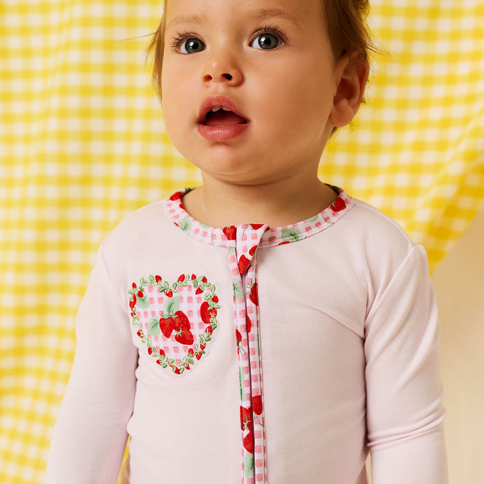 close up of baby wearing a bamboo viscose one piece with solid light pink fabric and strawberry printed side applique, standing on a white and yellow background